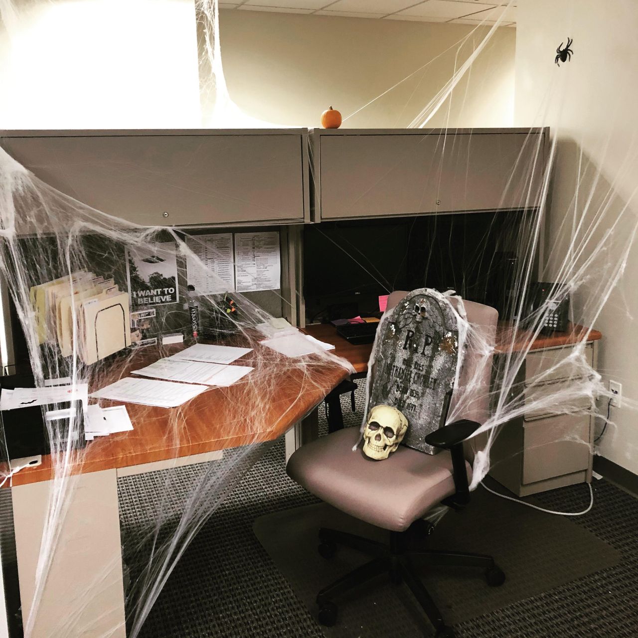Office cubicle covered in spider web, colleagues’ chair with a tombstone and topped with an adorable little pumpkin